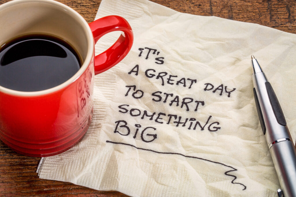 A red coffee mug sits on a napkin, as does a black pen. The words "It's a great day to start something BIG" appear in black ink on the napkin.