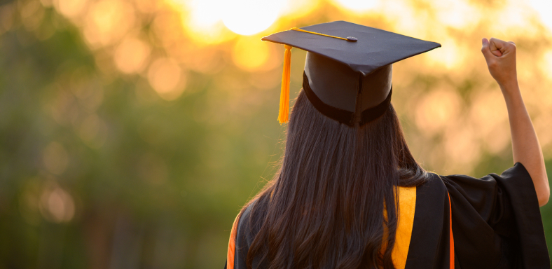 A photo from behind of a female graduate in cap and gown, her right fist proudly raised in the air.