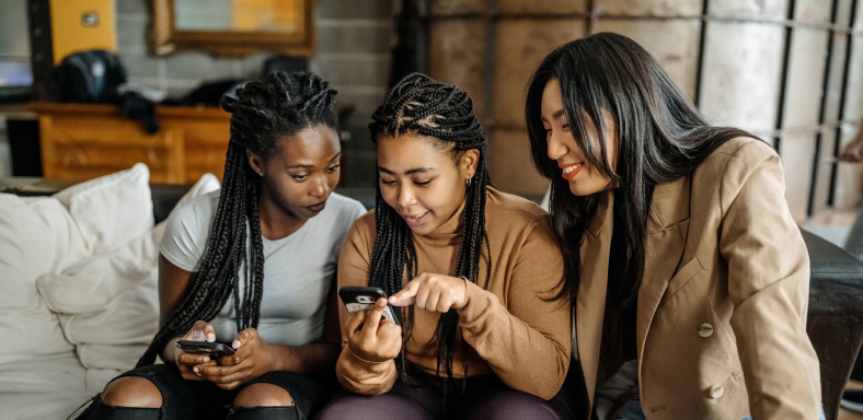 Three young women of color engaged in conversation about information on one of their smart phones.
