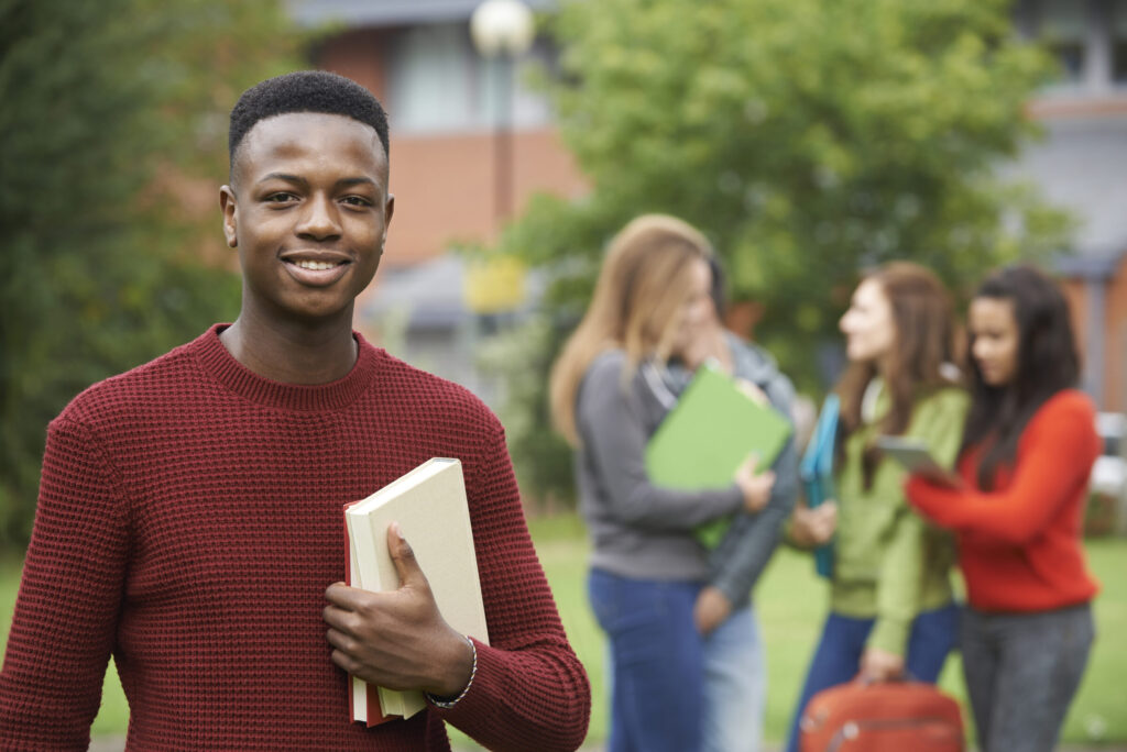 A young male student poses in the foreground while a group of female students discuss something in the background.